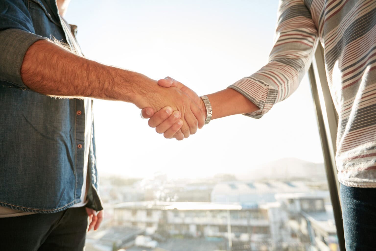 Two men shaking hands beside a sunlit window overlooking the city