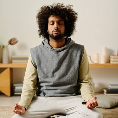 Young serene man in casualwear sitting in pose of lotus on the floor of bedroom and keeping eyes closed while practicing yoga exercise